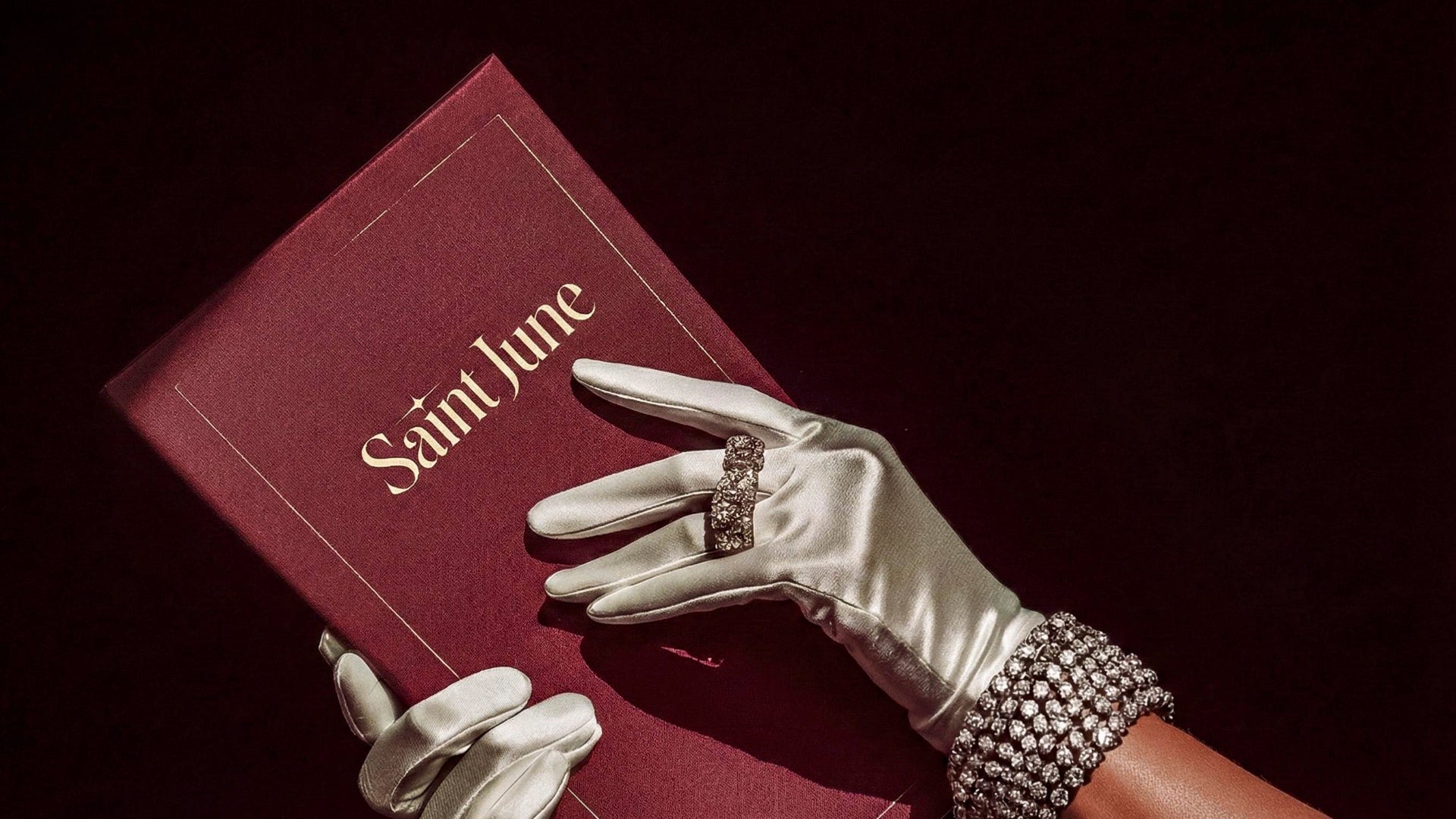 white gloved hands with jewellery opening a red book with the title saint june